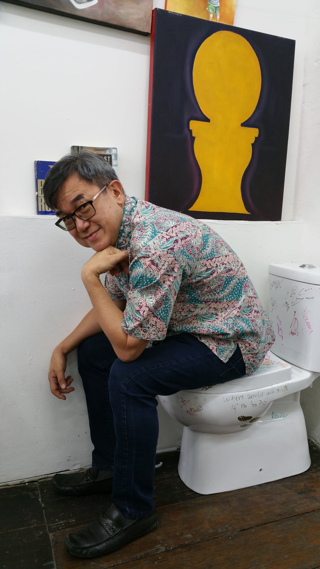 Jack Sim sitting fully clothed on a toilet, in a playful thinker’s pose